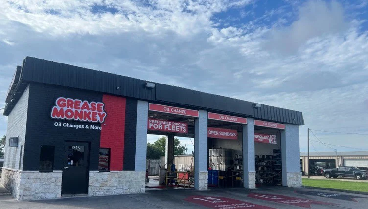 A Grease Monkey oil change service center with three open service bays and signage indicating oil changes, fleet pricing, and Sunday hours under a partly cloudy sky.